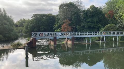 A weir with a walkway over it is reflected in the water. Behind it are trees and the river heading downstream