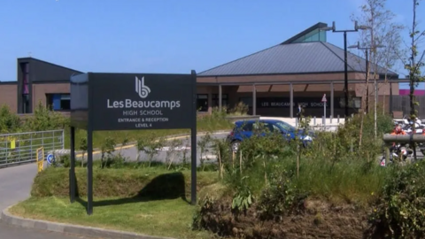 Les Beaucamps High School has been photographed showing a dark grey sign that reads Les Beaucamps High School. Behind the sign is the school with a light brown exterior and a light grey roof. 