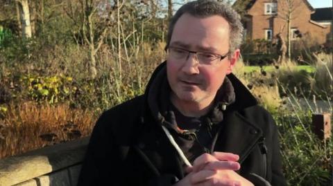 a man sits on a wooden bench in a garden during a BBC interview. He has short grey hair and is wearing glasses and a black winter coat. His hands are clasped together and he is looking downwards, as if thinking about something serious.