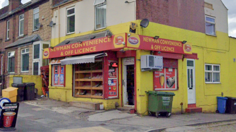 A bright yellow corner convenience shop called “Newman Convenience & Off Licence” with red signage and a window display, sits beside attached brick houses. Bins, a yellow container and small advertising boards stand outside on the pavement.