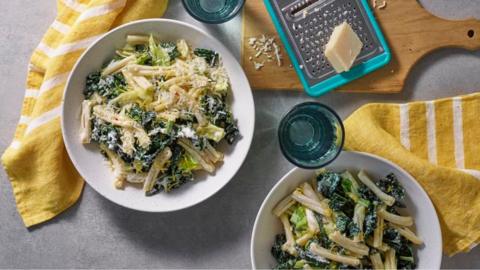 Two white bowls filled with pasta a green leaves, with yellow cloths, a cheese grater and chunk of parmesan 