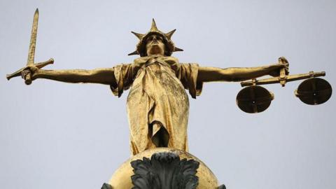 A statue of the gold scales of justice stands above the Old Bailey. The statue is holding a sword in one hand and scales in the other.