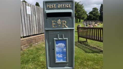 A dark green post box sits in a cemetery. It has a sign on the front with 'letters to heaven' against a blue sky and clouds.