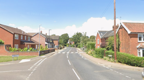 A residential street with a broken white line down the middle of the road. It is a sunny day with a blue sky. Red brick houses line either side of the street. A pedestrian crossing is marked on the road and junctions go off to the left and right in the foreground.