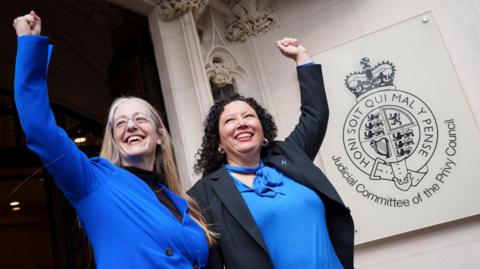 Campaigners Helen Joyce (left) and Maya Forstater (right) celebrate outside the UK Supreme Court in London. Helen has long blonde hair and glasses and is wearing a blue jacket and black top. Maya has curly dark hair and wears a blue blouse and black blazer.