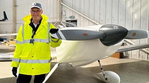 Man wearing a high viz jacket and grey cap standing next to a white plane with grey propeller