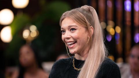 Becky Hill smiles with a blurred lit background behind her. She is wearing a black crew neck jumper and a gold necklace.