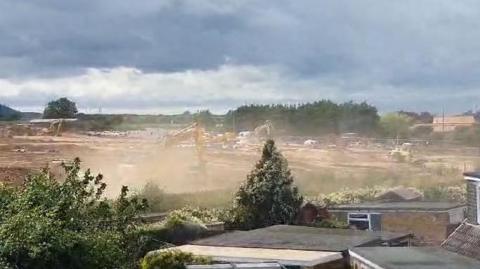 An overview of a building site with a digger partially obscured by a cloud of dust, with rooftop of nearby houses in the foreground.