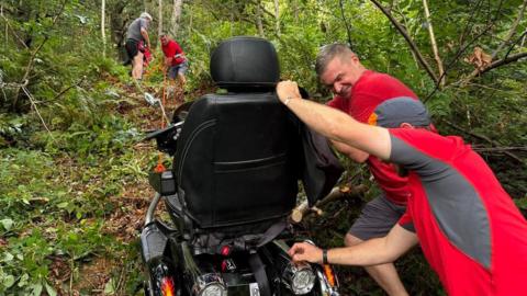 The mountain rescue team trying to recover the scooter. Two men are pushing it from behind up the hill. They are hearing red and grey t-shirts. At the top of the hill two men can be seen pulling the scooter up using orange rope.