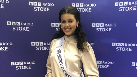 A woman wearing a pale-coloured top and a white sash is standing against a purple BBC Radio Stoke backdrop. She has curly black hair and is smiling for the photograph.