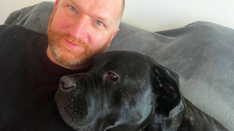 James Strauch with dog Bruno. James has a ginger beard.
Bruno is a large, black dog and he is resting on James' shoulder.