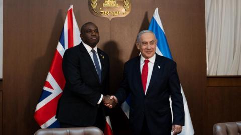 British Foreign Secretary David Lammy shakes hands with Israeli Prime Minister Benjamin Netanyahu in front of the flags of their respective countries. 