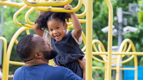 The image shows a father holding his a smiling child up as she holds onto a yellow climbing frame. The rest of the yellow structure can be seen in the background, along with trees which are blurred out in the image.