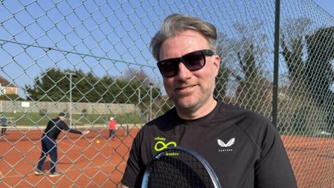 A man with greying hair wearing a black t-shirt and sunglasses is holding a tennis racquet and standing next to the fence of a tennis court. Behind him, people are playing tennis. 