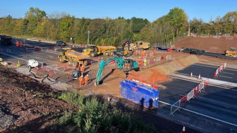 Large construction vehicles are beside a pile of rubble across the M4 motorway, which is fully closed. A number of contractors in orange high-vis clothing and white helmets are also visible. 