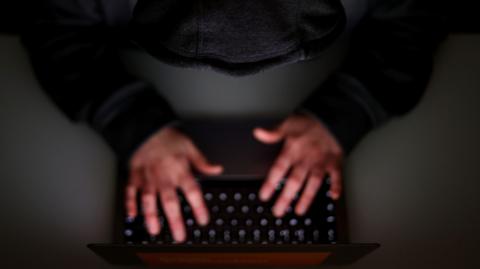 A stock image taken from a birds-eye view showing a young person in a grey hoodie typing on a laptop on a desk