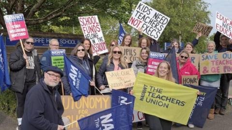 A group of 17 people holding large flags and banners on a picket line outside Corfe Hills School.