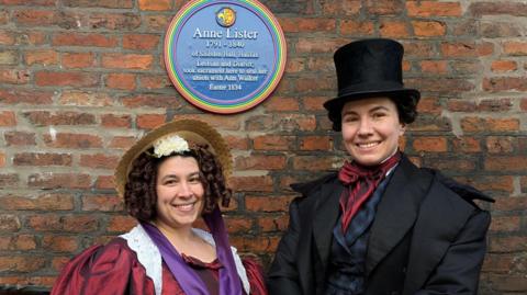 Two women, who are dressed up as Ann Walker and Anne Lister, are smiling to the camera in front of a rainbow plaque honoring Anne Lister.