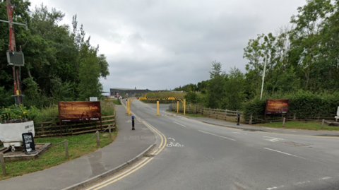 The entrance to Gedling Country Park, with trees and bushes either side of a single-carriageway road. Yellow barriers mark the entrance to the park