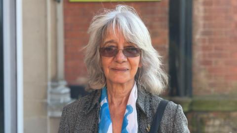 A profile image of Miriam Gifford, a 70-year-old woman with short grey hair and a fringe. She is wearing a white and sky-blue printed scarf, a grey jacket and glasses with a slightly tinted lens. 