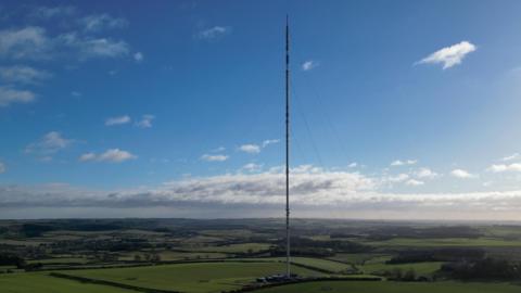 The Belmont transmitter - a tall, thin transmission mast rises from the middle of bright green fields, supported by fine guy wires. Under a clear blue sky with scattered clouds, the mast stands prominently above the open farmland, with small buildings at its base and low hills in the distance.