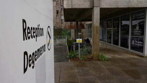 Outside of the Cornwall Council headquarters in Truro. A sign pointing towards the reception area is in the foreground. A person is walking along a path towards the building's entrance.