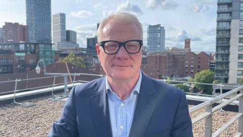 A man with short blond to grey hair is pictured standing on a rooftop area with a view overlooking Birmingham. He has thick black glasses on and his wearing a light blue shirt and navy blue jacket.