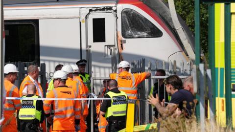 A large crowd of police and railway workers attending a damaged white train carriage.