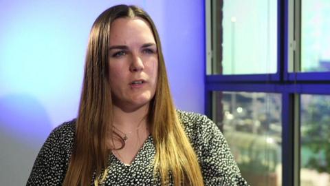 Charlene has long mid brown hair which she wears down. She is sat next to a window and is back lit with a blue/purple light. She is wearing a black and white top.