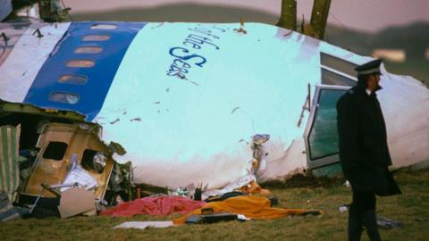 A police offer stands next to the wrecked cockpit of a passenger jet. It is lying on its side in a field.