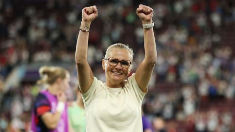 Sarina Wiegman raises her arms in the air and smiles after England's semi-final victory on 22 July 2025 in Geneva, Switzerland.