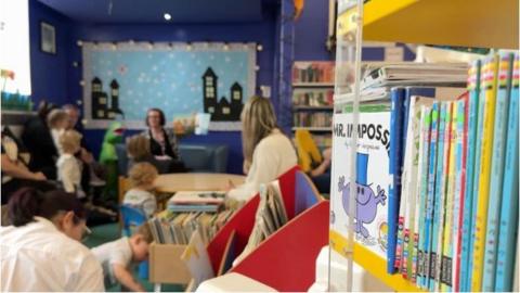 A story time session at the family library, you can see a bookshelf in the foreground and children and parents in the background listening to a woman read a story. She holds a book up.
