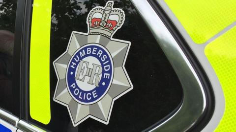 The rear side window of a yellow and blue Humberside Police car with the force's badge on the glass