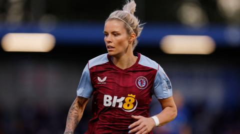A woman playing football while wearing a claret and blue Aston Villa shirt. Her blonde hair is tied back and she is looking to the right with her left arm partially raised. She has tattoos on her right arm.