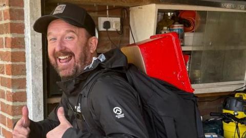 A laughing Stu Skinner gives a double thumbs up to the camera. On his back is a backpack with a red rectangular post box poking out the top. He has a full brown beard and is standing side-on to the camera. He appears to be in a domestic garage and is wearing black hiking gear and a black baseball cap. 