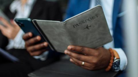 A close up of the hands of a person holding a booklet which says "passport" on the cover in one hand and a mobile phone in the other.