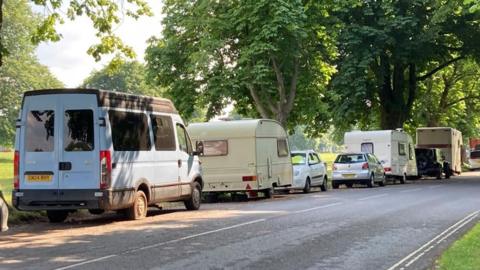 A row of vans, caravans, and cars are parked up on a street with a green field lined with trees.