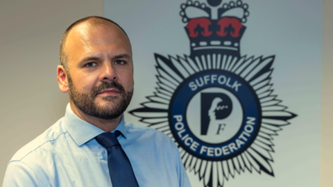 A head and shoulders image of Matt Paisley. He is wearing a light blue shirt and a navy tie. He is standing in front of a large Suffolk Police Federation emblem, which is on the white wall behind him. He is looking into the camera. 