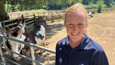 Hannah, a woman, wearing a blue T-shirt, smiling for the camera, while standing in front of a donkey enclosure.