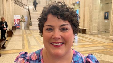 Maria Thomasson is standing in the middle of the Great Hall at Stormont. She is a young woman, with short, brown curly hair and she is wearing red lipstick and a pink and blue floral pattern dress. She is smiling at the camera.