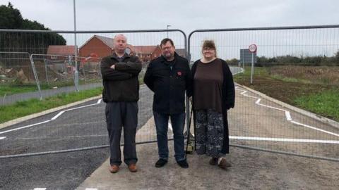 three people standing in front of fencing with new road and housing development in the background.