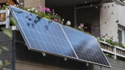 A solar panel hanging on a balcony, which also contains plants and flowers. 