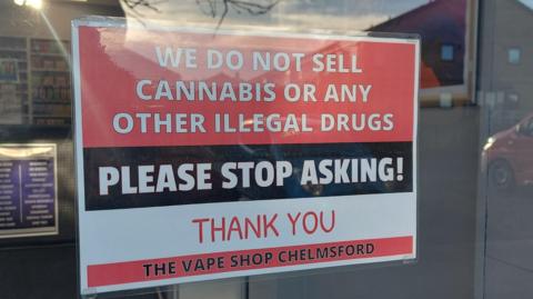 A sign behind a shop glass that says "we do not sell cannabis or any other illegal drugs, please stop asking, thank you, the vape shop, Chelmsford"