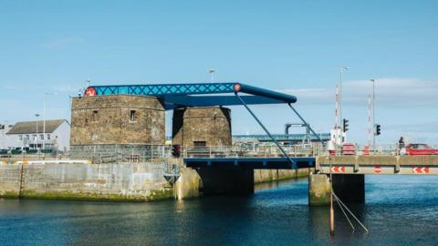 The lifting bridge in Douglas Harbour, which has two stone built towers beneath the blue lifting mechanism attached to the bridge, which is also blue. There is a red car travelling towards the raised barriers on the structure and the sea below is blue.