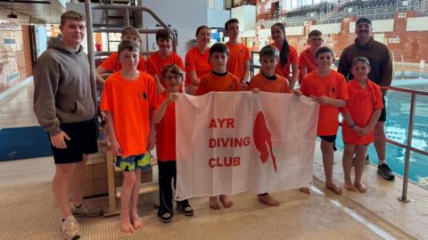 A group of kids wearing orange t-shirts stand by a swimming pool holding a white banner with red block capitals that read - Ayr Diving Club. Two adults stand at either side of the group wearing a dark coloured hoodie and smiling.
