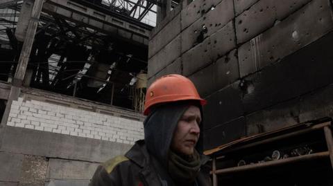 A workman in an orange safety helmet walks through a large charred industrial plant.