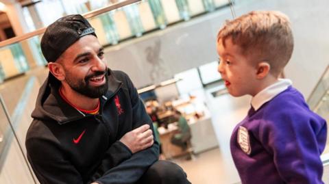 Mo Salah, with a trimmed black beard and who is wearing a baseball cap with the peak turned backwards and black zip-up Liverpool FC branded jacket, crouches down to speak to an eight-year-old boy who has light brown hair and a blue school jumper.