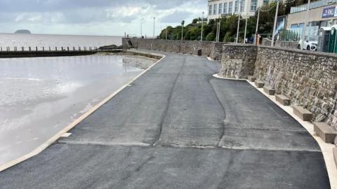 A dark grey, tarmac-like surface runs along the side of the marine lake, with a white edge and a stone wall on the other side. You can see the sea beyond the edge of the lake