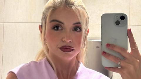A blonde girl with dark lipstick holding a phone taking a mirror selfie