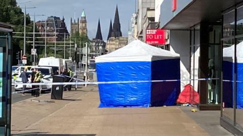 A blue and white police tent behind a corden on a pavement with police officers standing next to a police car